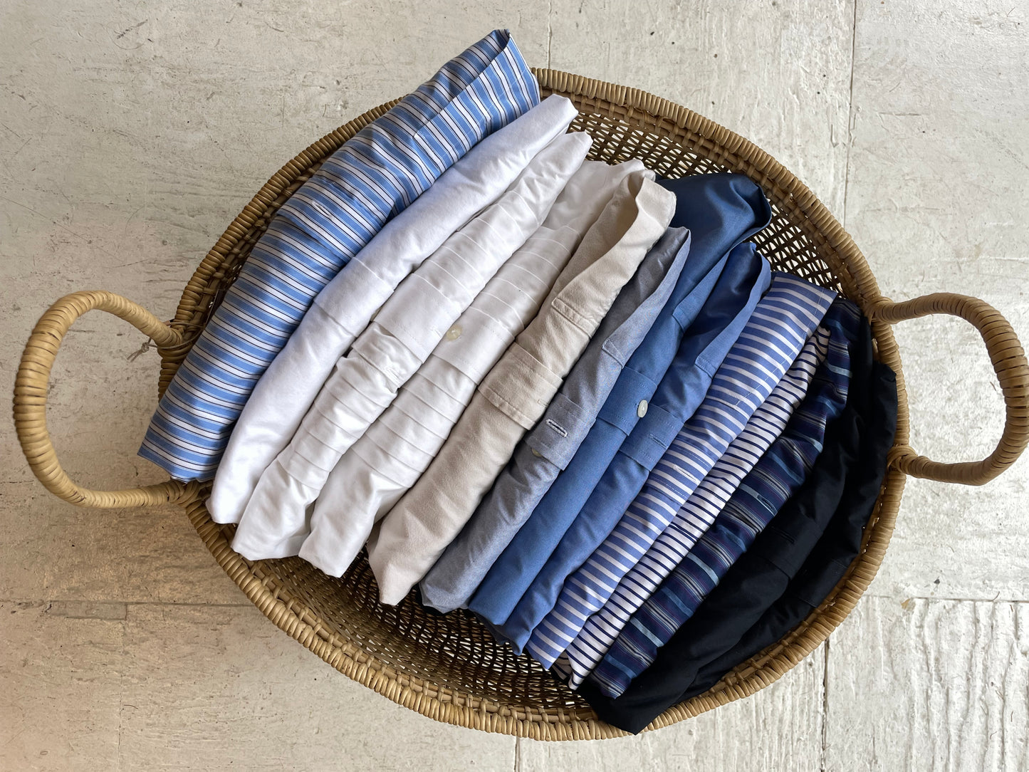 A collection of folded shirts in various shades of blue and white, neatly arranged in a wicker basket.